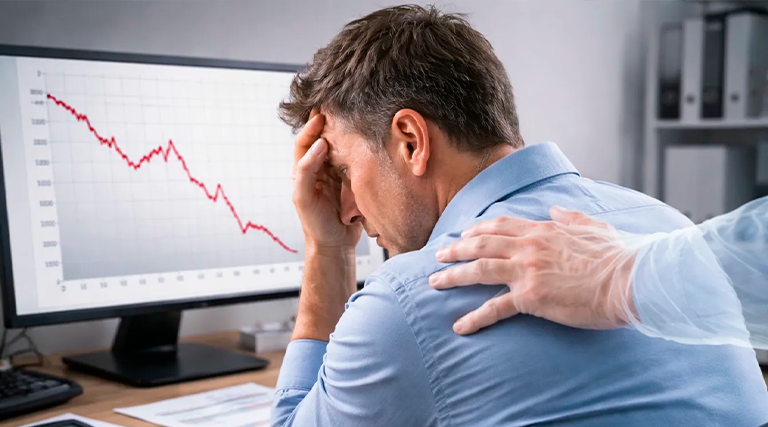 Homem com a cabeça apoiada nas mãos em frente ao computador com gráfico de queda em vermelho representando prejuízo financeiro enquanto outra pessoa coloca a mão em seu ombro em gesto de apoio, ilustrando a necessidade de um advogado para quem perdeu dinheiro em investimento