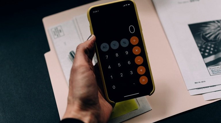 Mão segura smartphone com calculadora aberta sobre documentos para cálculo de lucros cessantes por atraso de imóvel