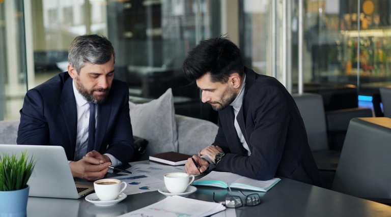 Braskem vai falir?: análise de documentos corporativos Braskem vai falir?: dois homens de terno analisando documentos e laptop em mesa de escritorio corporativo