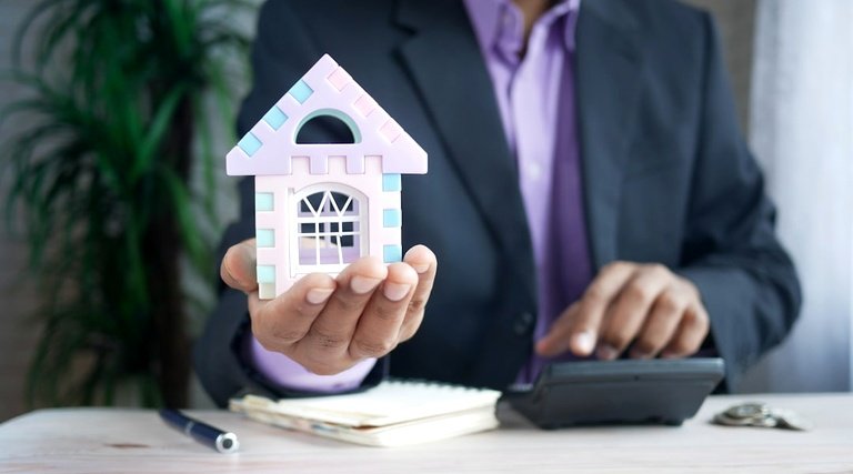 Atraso Entrega Imóvel Miniatura Mesa Profissional Atraso entrega imovel na planta: pessoa de terno segurando miniatura de casa com calculadora e documentos na mesa