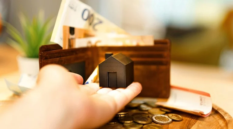 holding patrimonial vantagens e desvantagens: Mão segura miniatura de casa sobre mesa com carteira, cédulas e moedas